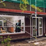 Een persoon die in een kamer staat en uit een groot raam kijkt in een bakstenen gebouw dat gerenoveerd wordt. Steigers omringen de buitenkant en bouwmaterialen liggen verspreid over de grond. Het interieur lijkt ingericht met verschillende items.