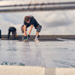 Twee bouwvakkers installeren isolatiematerialen op een dak. Eén arbeider, met kniebeschermers en een gereedschap, richt zich op het vastzetten van het materiaal terwijl de ander extra secties voorbereidt. Het beeld wordt omlijst door de sporten van een ladder.