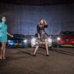 Drie vrouwen poseren dramatisch voor twee geparkeerde auto's met felle koplampen. Een draagt een medisch pak met een masker, een ander is gekleed in een zwart leren pak en de derde voert een handstand uit, gekleed in een zilveren bodysuit.