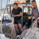 Twee vissers werken op een boot. De een maakt visnetten schoon en sorteert ze terwijl hij zwarte handschoenen draagt, en de ander houdt de netten dicht bij een stapel netten en een geruite doek. Ze worden omringd door visgerei en de boot ligt aangemeerd op een schilderachtige locatie.