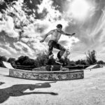 Een shirtloze skateboarder met een pet en camouflagebroek voert een truc uit in de lucht in een skatepark onder een zonnige, gedeeltelijk bewolkte lucht. De zwart-witfoto heeft een dynamische hoek die zijn schaduw op de grond vastlegt.