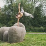 Een man zonder shirt voert een indrukwekkende parkourbeweging uit, balancerend op één hand op een grote ronde steen in een stadspark. Zijn lichaam is uitgestrekt in de lucht met één been omhoog, omringd door groen en bomen met een appartementengebouw op de achtergrond.