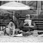 Een persoon bespeelt een panfluit terwijl hij op een deken op de grond zit. Ze staan naast een busje, met een parasol die schaduw biedt. Er staat een rolstoel naast hen en er liggen verschillende spullen verspreid over de deken.