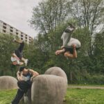 Drie mensen voeren parkour-stunts uit in een buitenpark. Eén persoon zit op een grote stenen constructie, terwijl een ander erboven in de lucht een salto maakt en de derde persoon een handstand uitvoert. Bomen en een woongebouw zijn op de achtergrond te zien.