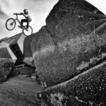 Een persoon met een helm voert een stunt uit door op een fiets over grote, oneffen rotsblokken te rijden. De zwart-witfoto legt de actie in de lucht vast, met de zon die een stervormig effect op de achtergrond creëert. De grond is zanderig en de lucht is overwegend helder.