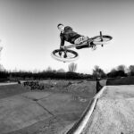 Een persoon voert een truc uit in de lucht op een BMX-fiets in een skatepark. De zwart-witfoto legt de rijder en de fiets vast die schuin boven de hellingen staan, met bomen en met graffiti bedekt beton zichtbaar op de achtergrond. De lucht is helder.