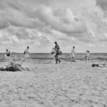 Zwart-witfoto van een strandtafereel. Meerdere mensen zijn verspreid over het zand, sommigen wandelen, anderen zitten op handdoeken en anderen spelen aan de waterkant. De lucht is gedeeltelijk bewolkt en de oceaan strekt zich uit tot aan de horizon op de achtergrond.