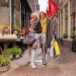 Twee vrouwen lopen arm in arm door een charmante, smalle straat vol winkels en bloemen, met verschillende boodschappentassen in hun handen. Beiden lachen, gekleed in casual, stijlvolle kleding met sjaals en jasjes, en lijken te genieten van een vrolijk dagje uit.