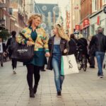 Een vrouw en een jong meisje lopen hand in hand door een levendige winkelstraat, elk met meerdere boodschappentassen. Winkelpuien met verschillende borden staan aan beide kanten van de drukke straat, en andere shoppers en voetgangers zijn zichtbaar op de achtergrond.