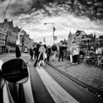 Zwart-witfoto van een drukke stadsstraat met voetgangers die lopen, geparkeerde fietsen en historische gebouwen onder een dramatische lucht. De foto is vervormd met een fisheye-effect, waardoor een gebogen perspectief van het stedelijke tafereel ontstaat.