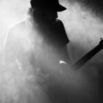 Een zwart-witfoto van een gitarist met een pet die een elektrische gitaar bespeelt, omgeven door dichte mist of rook op het podium. Het gezicht van de muzikant wordt verduisterd door schaduwen en mist, waardoor een mysterieuze en sfeervolle scène ontstaat.