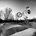 Een grijswaardenafbeelding van een persoon die een luchttruc uitvoert op een BMX-fiets in een skatepark. De rijder is halverwege de sprong, met de fiets gekanteld en beide wielen van de grond. Kale bomen op de achtergrond suggereren dat het winter is. Het skatepark heeft graffiti op de hellingen.