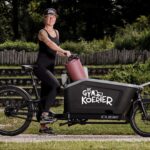 Een vrouw in zwarte sportkleding en een pet lacht terwijl ze naast een bakfiets staat. De fiets heeft een grote rechthoekige bak met het opschrift "De Gymkoerier" en bevat een roze cilindervormig object. Ze staat op een grindpad met groen en een houten hek op de achtergrond.