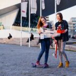 Twee jonge vrouwen staan buiten een modern gebouw. Beiden houden kaarten vast, de een op een skateboard en de ander met een skateboard naast zich. Ze lachen en zijn casual gekleed, terwijl er op de achtergrond vlaggen achter hen wapperen.