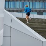 Een persoon in een blauw jasje en een zwarte short rent een brede buitentrap op. De achtergrond bevat een modern gebouw met grote ramen en reflecterende oppervlakken.