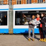 Drie mensen staan voor een blauw-witte tram. Eén persoon wijst vooruit terwijl hij een kaart vasthoudt. De andere twee houden camera's vast en kijken in de richting waarin gewezen wordt. Het lijken allemaal toeristen te zijn.