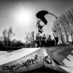 Een skateboarder voert in de lucht een truc uit op een ramp in een skatepark. Meerdere mensen zitten op de rand van de ramp en kijken toe. De zon schijnt fel aan een heldere hemel met bomen op de achtergrond. De ramp is bedekt met graffiti. Zwart-witfoto.