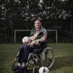 Een oudere man in een rolstoel houdt een voetbal op zijn schoot, met een andere voetbal op de grond naast hem. Hij is gewikkeld in een groen-witte sjaal, zittend op een grasveld met een voetbaldoel op de achtergrond. Bewolkte luchten en bomen omlijsten het tafereel.