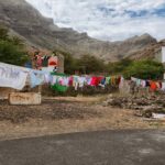 Kleding in verschillende kleuren hangt aan een waslijn die tussen stenen muren is gespannen in een buitenomgeving. De achtergrond bestaat uit hoge, ruige bergen onder een gedeeltelijk bewolkte lucht en bescheiden stenen gebouwen. De grond is een mix van aarde, rotsen en schaarse vegetatie.