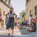 Een groep toeristen loopt op een zonnige dag door een smalle, geplaveide straat tussen historische stenen gebouwen. Een vrouw in een jurk staat op de voorgrond met haar handen in haar zij, en een kind zit op de grond rechts, leunend tegen een muur.