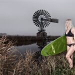 Een persoon in een zwarte bikini houdt een groene surfplank vast en staat bij een watermassa met een windmolen op de achtergrond. De lucht is bewolkt en het gras om hen heen is hoog en bruin. Het tafereel lijkt landelijk en sereen.