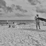 Zwart-witfoto van een strandtafereel. Een persoon rechts schudt een deken uit, een ander persoon zit en kijkt naar een tas in het midden, en een derde loopt langs de kust op de achtergrond. De lucht is gedeeltelijk bewolkt en de zee is kalm.