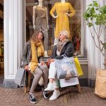 Twee vrouwen zitten op een bankje buiten een kledingwinkel, lachend en met boodschappentassen in hun handen. Beiden dragen sjaals en casual kleding. Mannequins die modieuze jurken tentoonstellen zijn zichtbaar in de winkel achter hen. Een kleine boom in een pot voegt groen toe aan het tafereel.