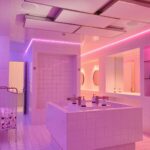 Een moderne badkamer met neonroze en paarse verlichting. De kamer heeft betegelde muren en vloeren, een centraal eiland met vier wastafels, spiegels en planken aan de muren. In de hoek staat een rek met meerdere rollen toiletpapier.