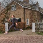 Een vrouw in zwarte sportkleding en kleurrijke hardloopschoenen wordt vastgelegd terwijl ze langs een bakstenen huis rent met groen en bomen eromheen. Ze lijkt geconcentreerd, met haar haar naar achteren gebonden en haar armen en benen in beweging.