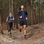 Twee mensen rennen op een pad door een bos. De persoon vooraan draagt een blauw shirt met lange mouwen, zwarte shorts en een zwarte hydratatierugzak, terwijl de persoon achter hen een blauw vest en zwarte leggings draagt. Bomen en een onverhard pad omringen hen.