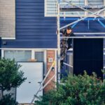 Een bouwvakker klimt op een steiger voor een modern blauw huis met witte afwerking. Groen gebladerte is zichtbaar op de voorgrond. De arbeider draagt veiligheidsuitrusting en lijkt midden in een taak te zitten.