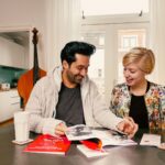 Een man en een vrouw zitten aan een tafel in een gezellig huis, bladerend door tijdschriften en pamfletten. De man draagt een grijs vest en lacht met de vrouw, die een glas vasthoudt. De kamer heeft een contrabas, een kleine tafellamp en lichtgekleurde gordijnen.