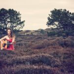 Een vrouw in een rode jurk zit in een veld met hoog gras en speelt op een akoestische gitaar. Twee grote bomen en een bewolkte lucht zijn op de achtergrond. De scène heeft een serene en enigszins grillige sfeer.