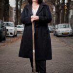 Een vrouw met krullend haar en een bril staat op een straat met bomen, met een schep in haar hand. Ze draagt een lange, donkere jas over een wit shirt met een donkere broek. Geparkeerde auto's staan aan beide kanten van de straat, met wat bladeren verspreid op de grond.