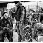 Een zwart-witfoto toont een oudere man die zit en een boekje vasthoudt, terwijl een jongere man in de buurt staat. Ze bevinden zich op een openluchtmarkt met tenten en kraampjes op de achtergrond, met een vintage motorfiets en verschillende items die worden tentoongesteld.