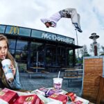 Een jonge vrouw nipt aan een drankje en eet frietjes aan een tafeltje buiten voor een McDonald's. Op de achtergrond voert iemand een indrukwekkende salto in de lucht uit, waarmee hij de aandacht trekt van omstanders in de buurt. Het McDonald's-bord en -logo zijn prominent aanwezig boven de ingang.