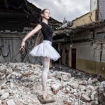 Een ballerina in een zwart maillot en een witte tutu poseert sierlijk op haar puntschoenen te midden van de ruïnes van een ingestort gebouw. Het contrast tussen haar elegantie en de verwoesting om haar heen is opvallend. De lucht is bewolkt, wat bijdraagt aan de sombere stemming.