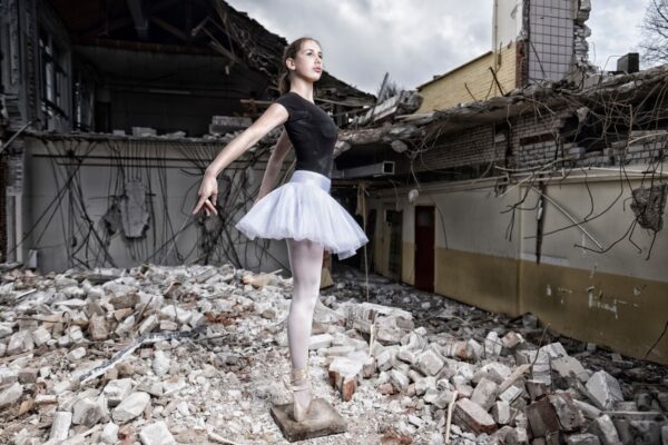 Een ballerina in een zwart maillot en een witte tutu poseert sierlijk op haar puntschoenen te midden van de ruïnes van een ingestort gebouw. Het contrast tussen haar elegantie en de verwoesting om haar heen is opvallend. De lucht is bewolkt, wat bijdraagt aan de sombere stemming.