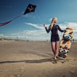 Een persoon in een wetsuit staat op een zandstrand met een vlieger in de ene hand en een kleurrijk kiteboard in de andere. De lucht is gedeeltelijk bewolkt met de oceaan op de achtergrond.