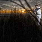 Een honkbalspeler staat in een batting stance met een knuppel, klaar om te slaan, omringd door hoog gras. Hij draagt een wit uniform en een zwarte pet. De lucht is geschilderd met dramatische oranje en grijze tinten, wat doet denken aan zonsondergang of zonsopgang.