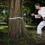 Een persoon die een witte martial arts gi draagt staat in een park en oefent bewegingen met een boom omwikkeld met een witte band. Het park heeft weelderig groen gras en hoge bomen, met zonlicht dat door het gebladerte heen filtert.