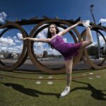 Een jonge kunstschaatsster voert een sierlijke pose uit op rolschaatsen, gekleed in een paarse jurk. Ze balanceert op één been, strekt het andere been en haar arm uit. De achtergrond toont grote metalen cirkels tegen een gedeeltelijk bewolkte lucht, met gebouwen en bomen in de verte.