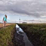 Een jonge vrouw in een turquoise shirt draagt een surfplank over een smal pad tussen twee velden. Op de achtergrond staan windturbines verspreid over het landschap onder een bewolkte lucht.