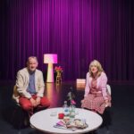 Een man en een vrouw zitten naast een ronde witte tafel op een podium met donkerpaarse gordijnen. Op de tafel liggen bloemen, tijdschriften en glazen. De man draagt een beige jas en een rode broek, en de vrouw draagt een roze en rood geblokte jurk. Een lamp en een klein tafeltje staan op de achtergrond.