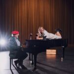 Een man met een rode hoed zit achter een vleugel en speelt muziek, terwijl een vrouw op de piano ligt en lacht met haar hand op haar kin. Twee wijnglazen en een wijnfles staan op de piano. De scène speelt zich af op een podium met een achtergrond van bruine gordijnen.