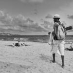 Een zwart-witfoto van een strandscène met een verkoper die over het zand loopt. De verkoper, met een hoed en rugzak, draagt artikelen die te koop zijn, hangend aan beide kanten. Op de achtergrond liggen mensen op het strand bij de oceaan onder een bewolkte lucht.