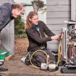 Een technicus knielt op de grond en onderzoekt een open warmtepompunit, waarbij hij een gereedschap op de bedrading gebruikt. Een andere persoon staat naast hem en kijkt aandachtig toe. Ze werken buiten bij een bakstenen muur en een houten schuur. Verschillende gereedschappen en apparatuur zijn om hen heen zichtbaar.