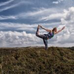 Een persoon voert een yogahouding uit die de dansershouding wordt genoemd op een met gras begroeide heuveltop onder een gedeeltelijk bewolkte lucht. De persoon balanceert op één been terwijl hij het andere been en beide armen uitstrekt om een sierlijke boog te vormen.