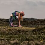 Een persoon voert een kameelhouding (Ustrasana) yogapositie uit op een grasveld met een heuvelachtig landschap op de achtergrond. Ze dragen kleurrijke yogakleding en hun rug is gebogen met hun handen rustend op hun hielen, omhoog kijkend. De lucht is bewolkt.