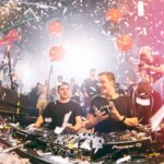 Twee dj's staan op het podium in een levendige, volle club. De sfeer is levendig met vallende confetti en ballonnen die door de lucht zweven. Eén dj houdt een koptelefoon vast, terwijl de ander de apparatuur aanpast. Clubgangers en fotografen omringen het verhoogde podium.